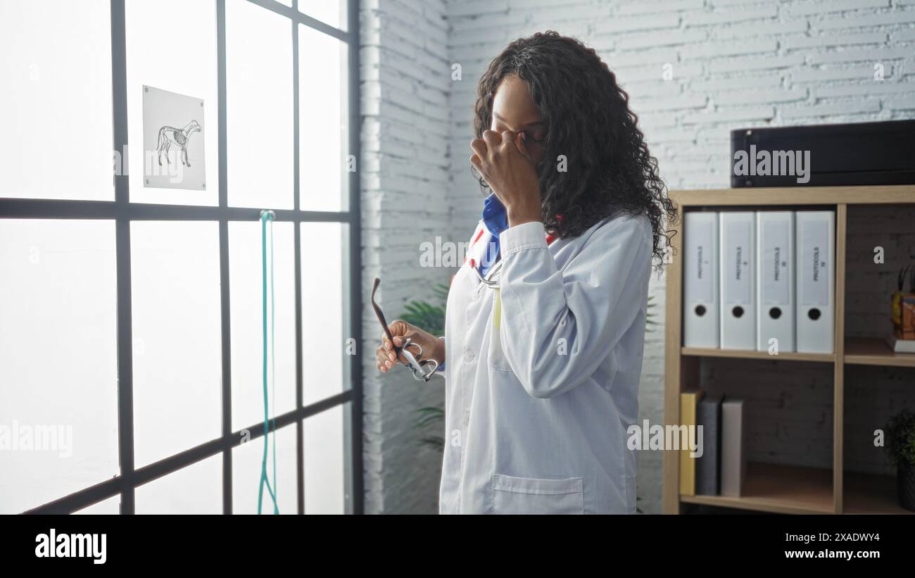 Una giovane donna afroamericana veterinaria con i capelli ricci in un camice da laboratorio bianco che le sfrega gli occhi stancamente in una stanza di clinica veterinaria al chiuso che tiene la g Foto Stock