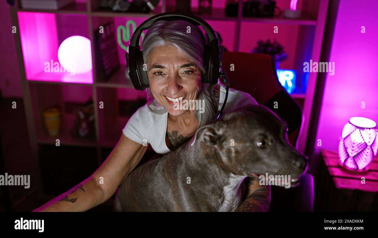 Una donna dai capelli grigi sorridente di mezza età, cuffie a dondolo e il suo cane in una vivace sala da gioco, che apprezza l'intrattenimento digitale Foto Stock