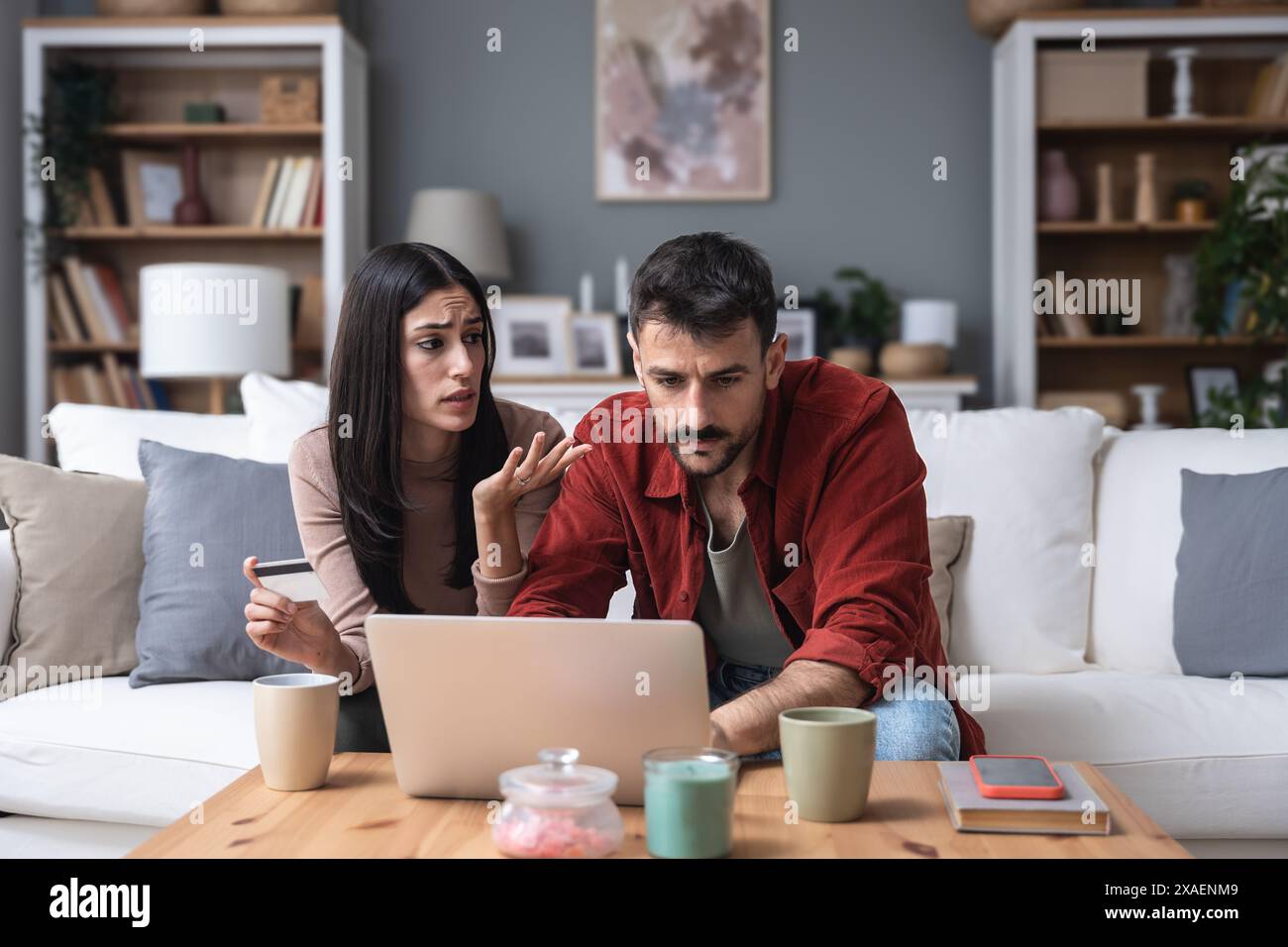 Una giovane coppia seduta a casa con un computer portatile che ha problemi con il limite della carta di credito e con le banche. Uomo e donna nervosi che cercano di trovare dove si trovano Foto Stock