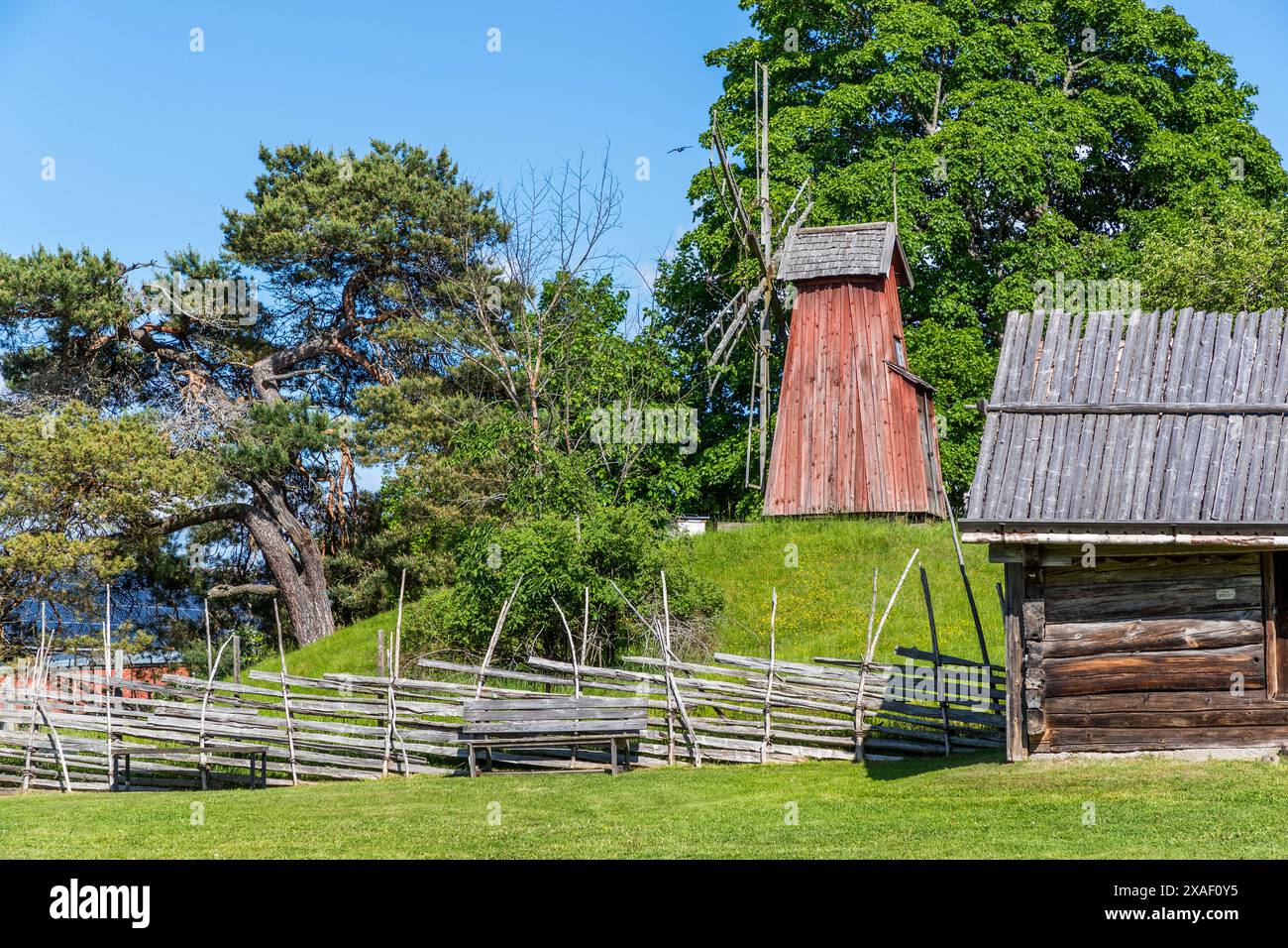 Un mulino a posta e una capanna di tronchi si trovano nel museo all'aperto Disagården vicino a Gamla Uppsala dietro una recinzione a rail diviso sinuosa, con una panchina in legno che invita i visitatori a riposarsi in primo piano. Gli edifici storici del museo risalgono tra il XVI e il XIX secolo e sono stati trasferiti in questo sito per formare il complesso all'aperto. Ärnavägen, Uppsala, Contea di Uppsala, Svezia. Foto Stock