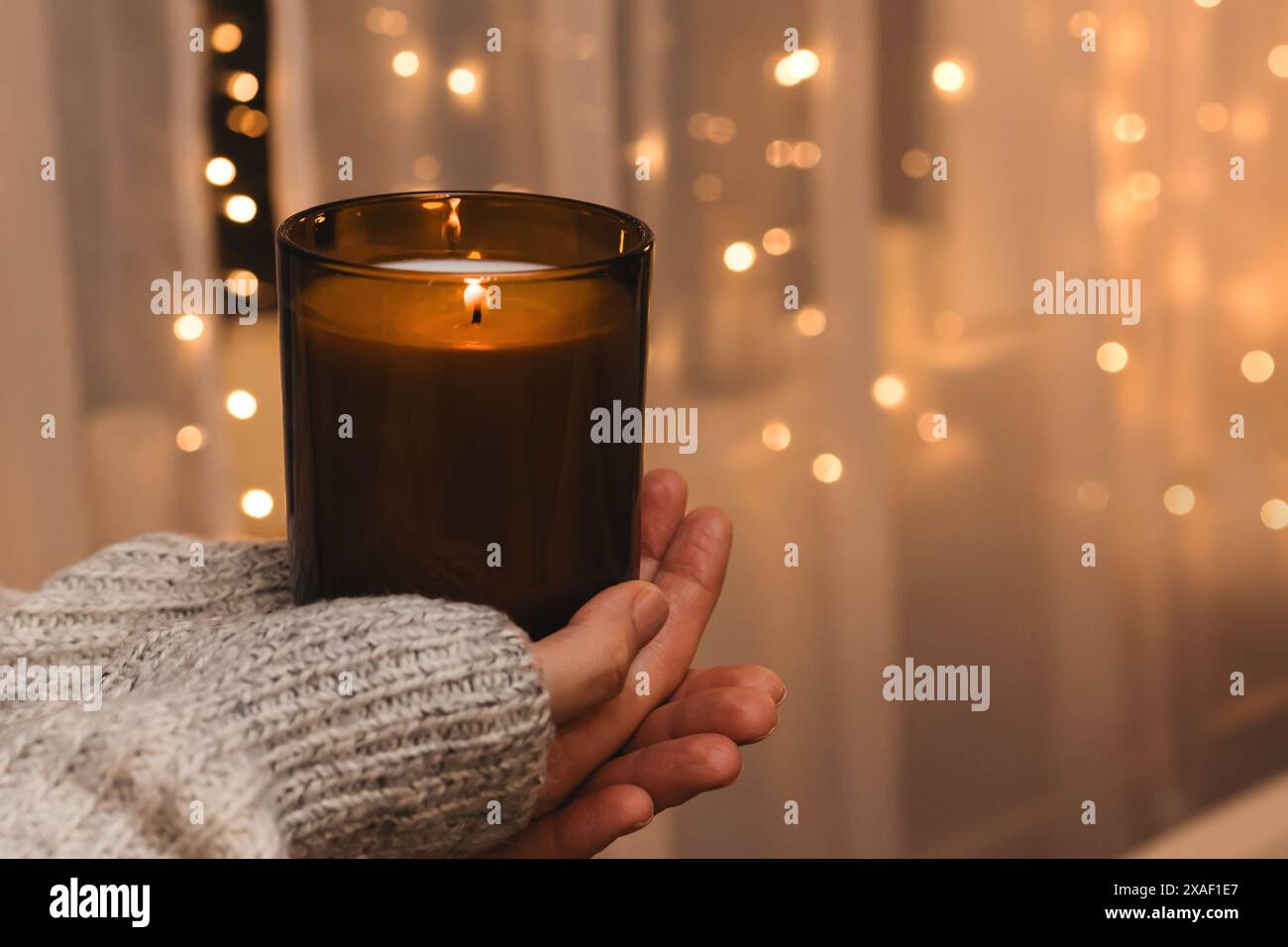 Donna con le mani che tiene in mano una candela accesa con luci sfocate sullo sfondo, un'atmosfera accogliente invernale Foto Stock