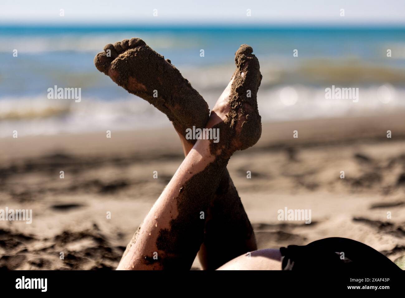 Primo piano di piedi di sabbia fangosa attraversati contro un oceano azzurro e una spiaggia soleggiata, che ritrae relax e vibrazioni estive. Foto Stock