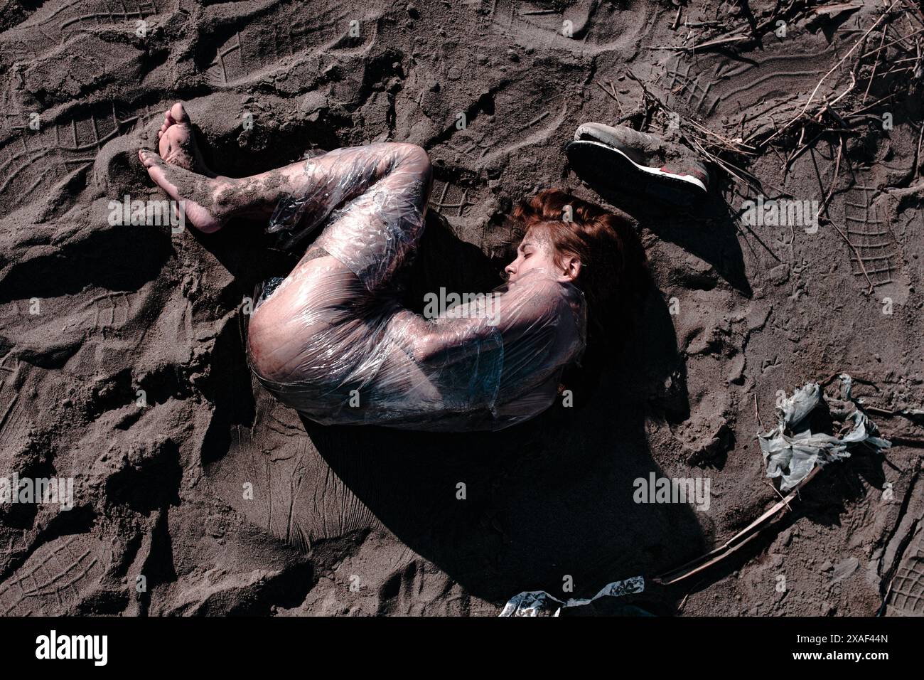 Immagine inquietante di una giovane donna avvolta in un involucro di plastica, sdraiata su una spiaggia sabbiosa, evocando temi ambientali e impatto umano. Foto Stock