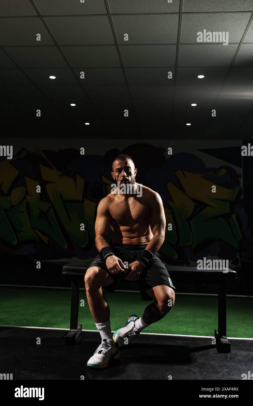 Atleta maschile concentrato e muscoloso si siede su una panchina e si prende una pausa in una palestra moderna con un vivace sfondo di graffiti. Acquisizione di un momento di determinatio Foto Stock
