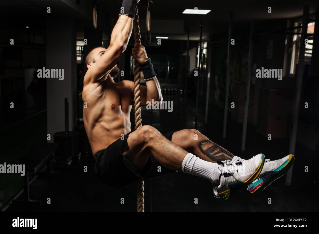 Un uomo muscoloso che sale su una corda in una palestra, mostrando forza, agilità e fitness. La scena cattura un momento di intenso sforzo fisico. Foto Stock