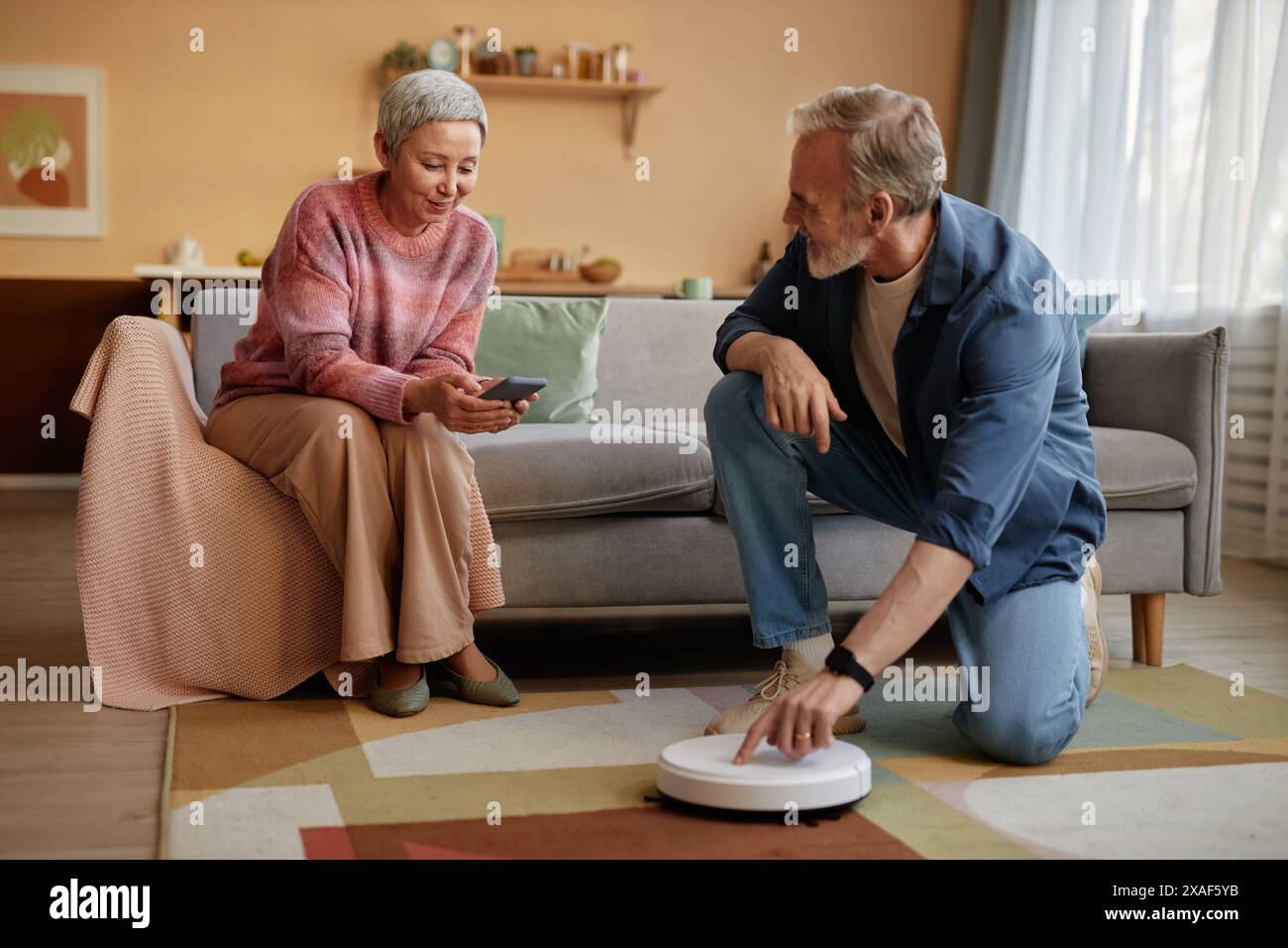 Ritratto di un anziano che preme il pulsante sull'aspirapolvere robot con una donna matura moderna che imposta i dispositivi domestici intelligenti tramite spazio di copia dello smartphone Foto Stock
