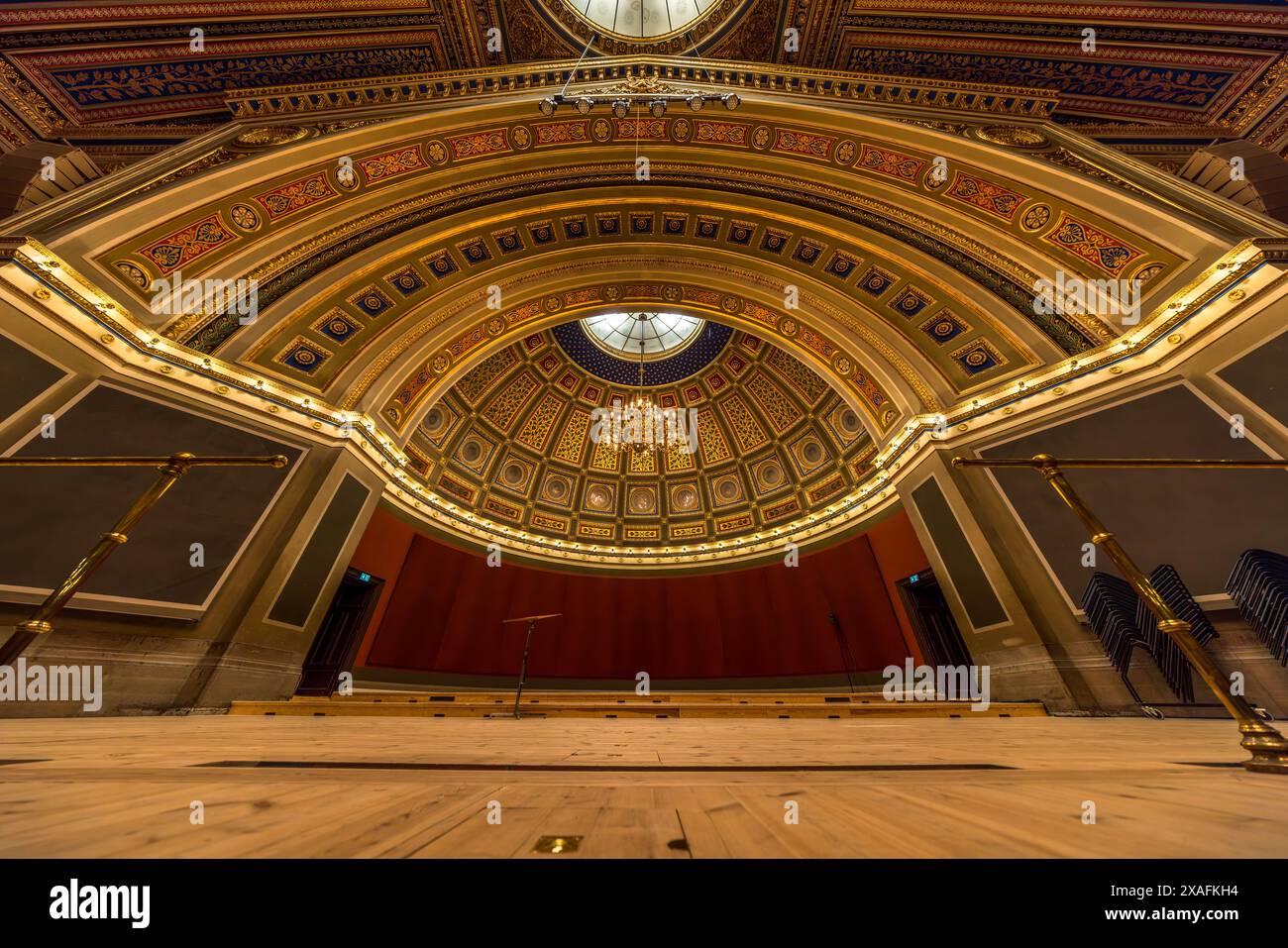 Un sipario rosso incornicia la vista dal palcoscenico fino alla cupola a cassettoni blu e oro del Grande Auditorium (Universitetsaulan) nell'edificio principale dell'Università di Uppsala (Universitetshuset). Un leggio si trova da solo sul pavimento di legno del palcoscenico, con sedie pieghevoli impilate appoggiate al muro a destra. La sala ha una capienza di circa 1.800 persone. L'architetto Herman Teodor Holmgren ha progettato l'edificio, completato nel 1887, in stile rinascimentale italiano. Biskopsgatan, Uppsala, Contea di Uppsala, Svezia. Foto Stock