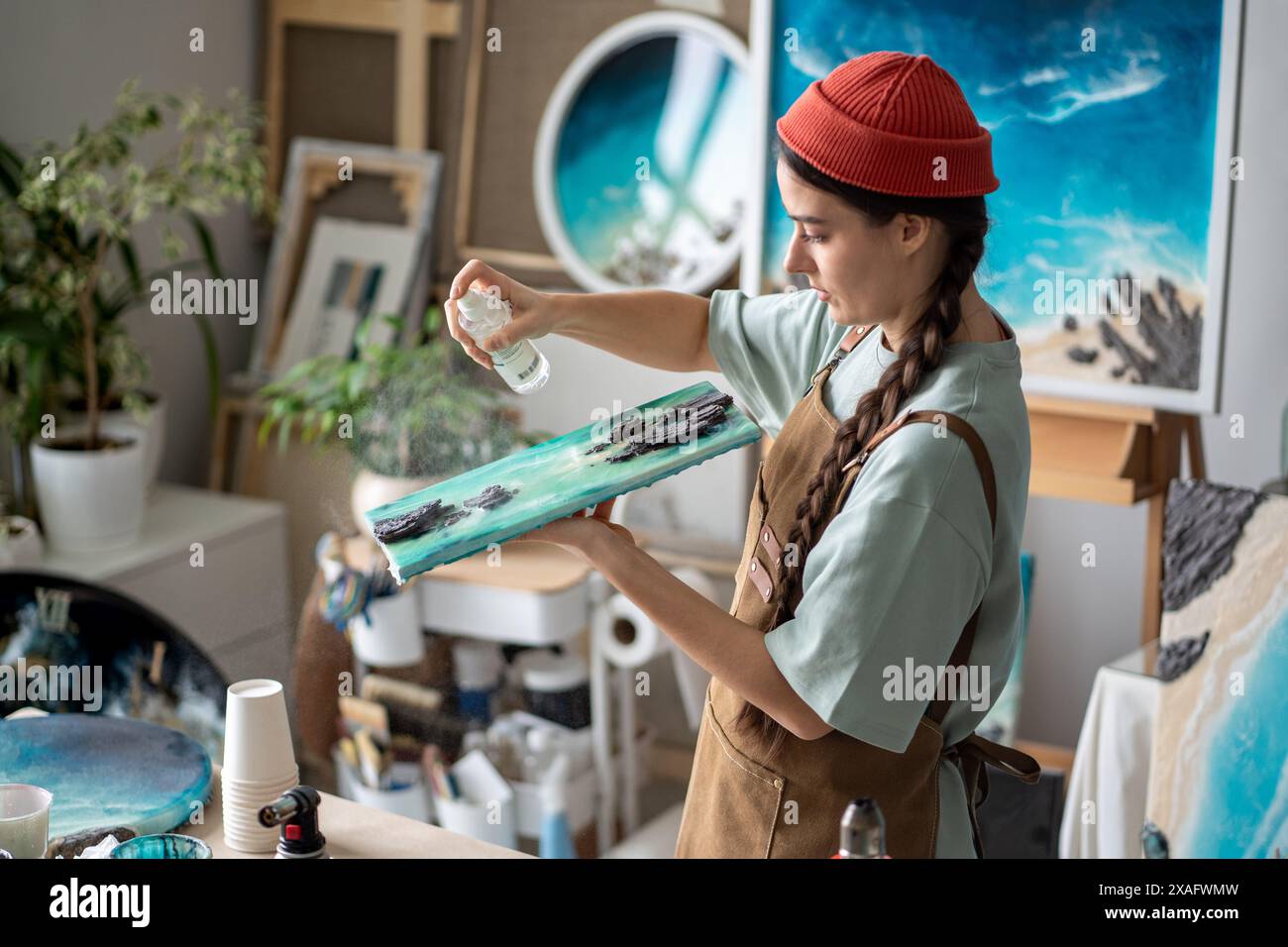 Artista donna incentrata che tiene tele, flacone spray che completa il lavoro finale sulla pittura in resina epossidica Foto Stock