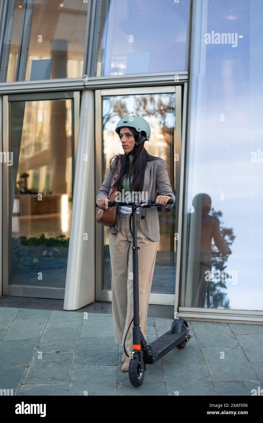 una donna d'affari verticale viene vista uscire da un edificio di uffici con uno scooter elettrico e un casco, mostrando il suo viaggio verso l'esterno nel rispetto dell'ambiente Foto Stock