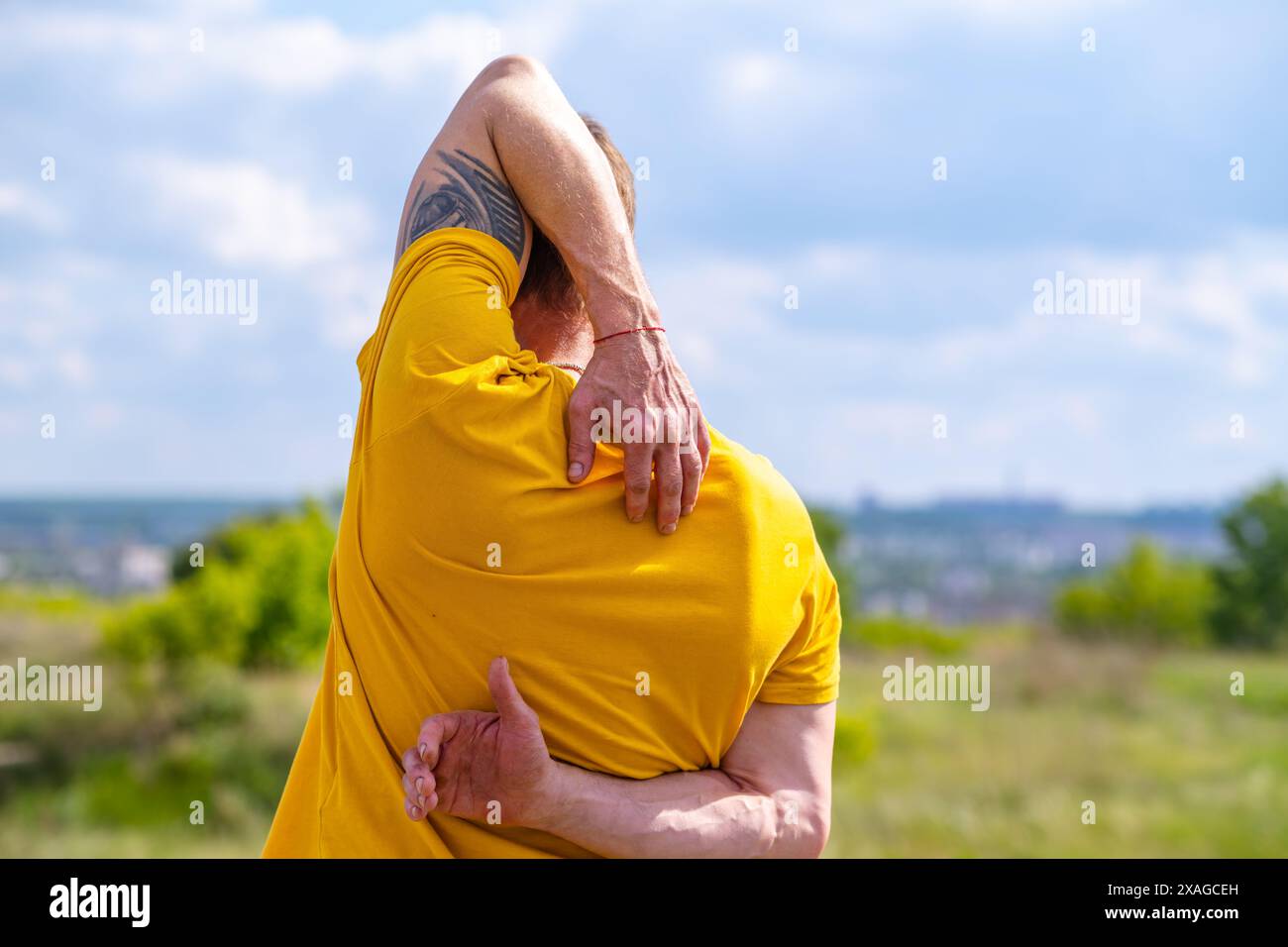 Un uomo con una t-shirt gialla si allunga il braccio mentre è in piedi fuori in una giornata di sole. Sta allungando i muscoli del braccio e della spalla. Foto Stock Un uomo con una t-shirt gialla si allunga il braccio mentre è in piedi fuori in una giornata di sole. Sta allungando i muscoli del braccio e della spalla. Foto Stock