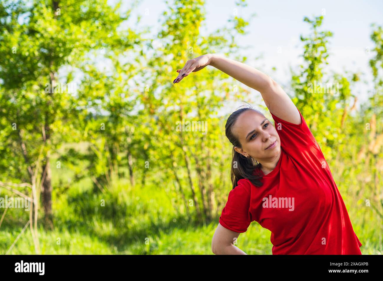 Donna che indossa una camicia rossa, che si allunga in un campo erboso. Ha il braccio destro esteso sopra la testa e la mano sinistra sull'anca Foto Stock