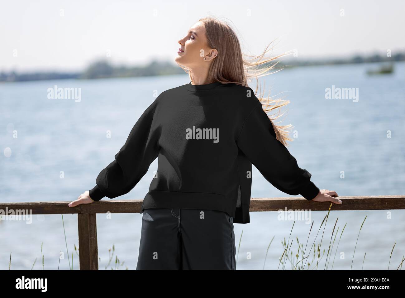 Mockup di una felpa nera su una ragazza sullo sfondo di un fiume, un molo, con il sole, vista frontale dell'abbigliamento. Modello per abiti eleganti da donna Foto Stock
