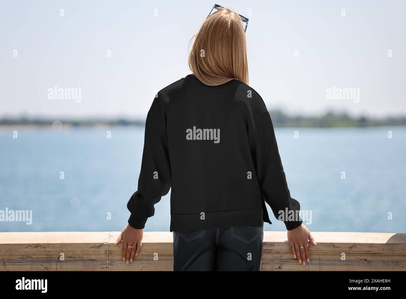 Modello di una felpa nera su una ragazza dai capelli chiari in condizioni di sole sul molo, sullo sfondo di un fiume, vista posteriore, per la pubblicità. Mockup di Foto Stock