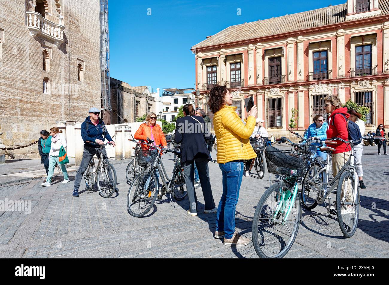 Un gruppo di ciclisti si riunisce in una soleggiata piazza di Siviglia, vicino agli edifici storici, mentre una persona scatta una foto e gli altri chiacchierano Foto Stock