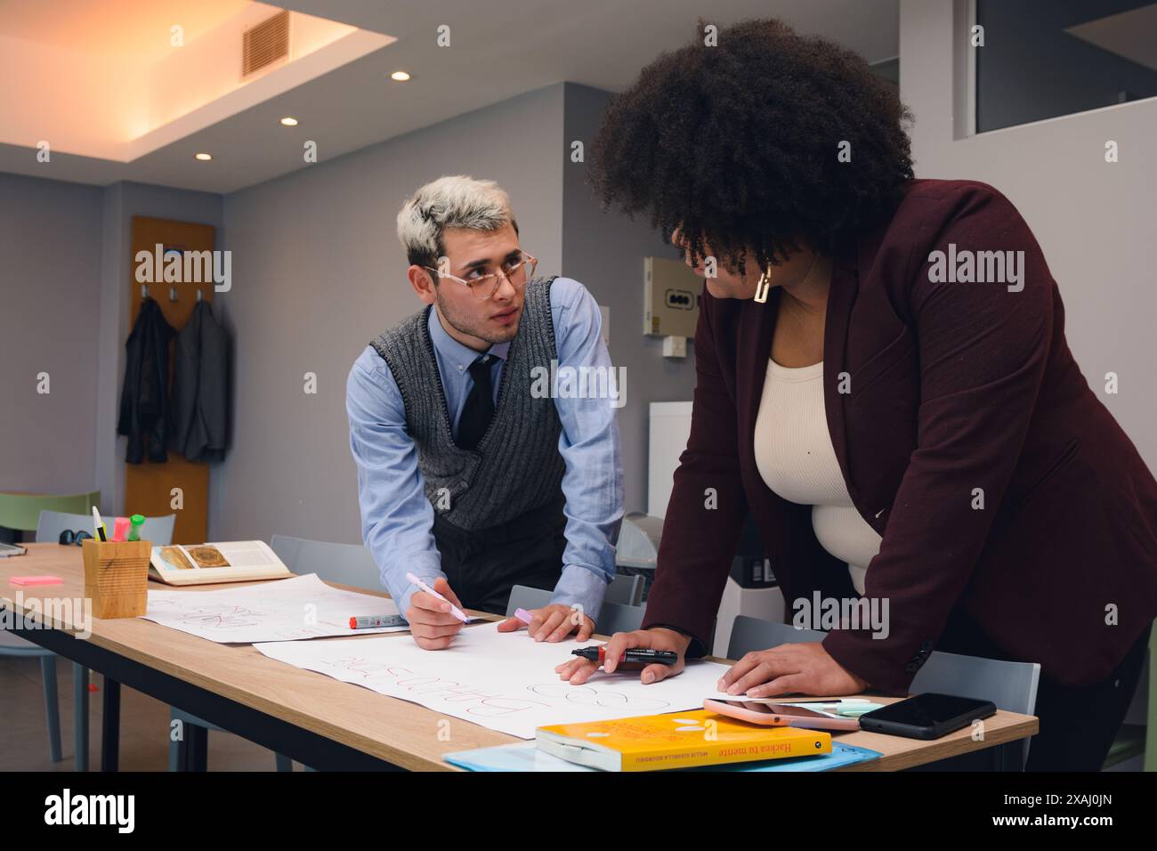 il giovane in ufficio parla con la sua collega donna dei documenti sul tavolo. Ha in mano una penna e vi mostra il percorso di lavoro che sta progettando Foto Stock