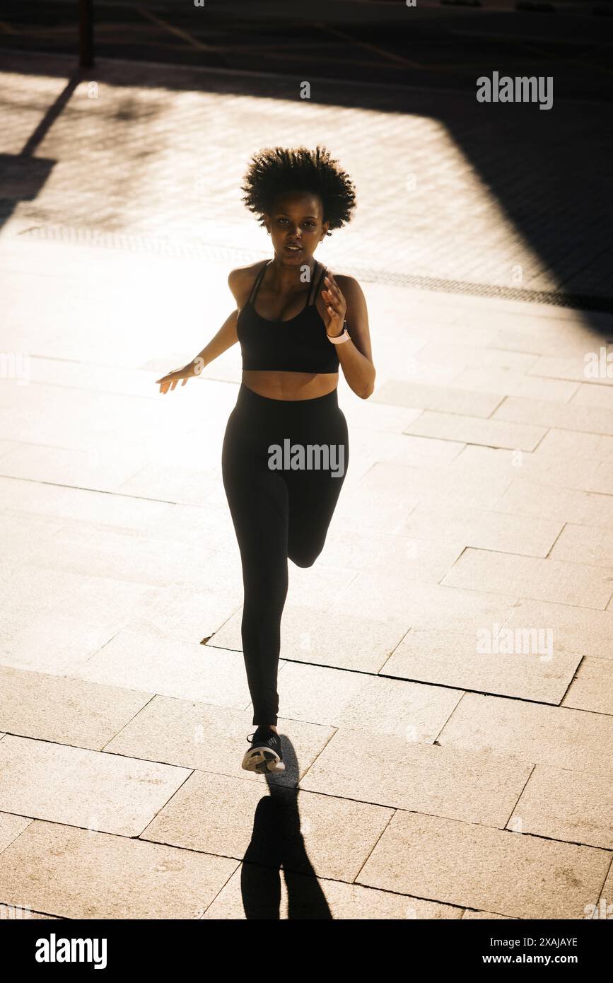 Donna che corre con determinazione su una strada urbana illuminata dal sole. Donna con una postura atletica che fa jogging su un set urbano. Foto Stock