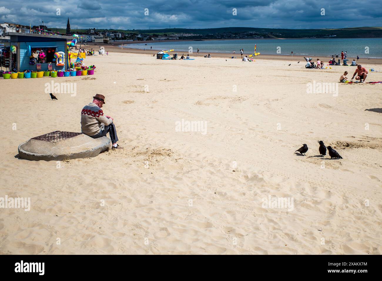 L'uomo solitario su una spiaggia quasi vuota con tre corvi che lo guardano. Spiaggia di Weymouth, Dorset. Solitudine, pensiero, comunicazione. Uccelli. Foto Stock