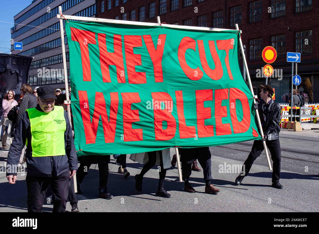 Ci tagliano l'emorragia. Persone che portano un grande striscione alla marcia del Labor Day nel quartiere Hakaniemi di Helsinki, Finlandia. Foto Stock
