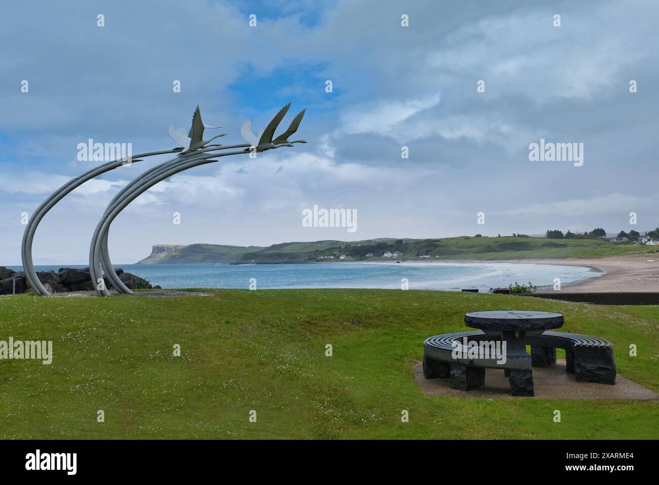 I bambini di Lir Swans scultura, una panchina circolare in pietra sul lungomare di Ballycastle, Irlanda del Nord, con mare blu, cielo blu e sfondo di montagne Foto Stock