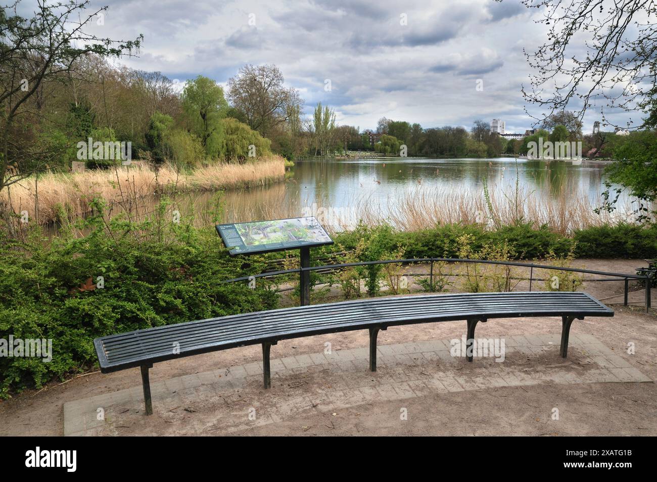 Una panchina vuota e un tabellone informativo/informativo accanto al Regent's Park Boating Lake in una calda giornata, a nord-ovest di Londra, Inghilterra, Regno Unito Foto Stock