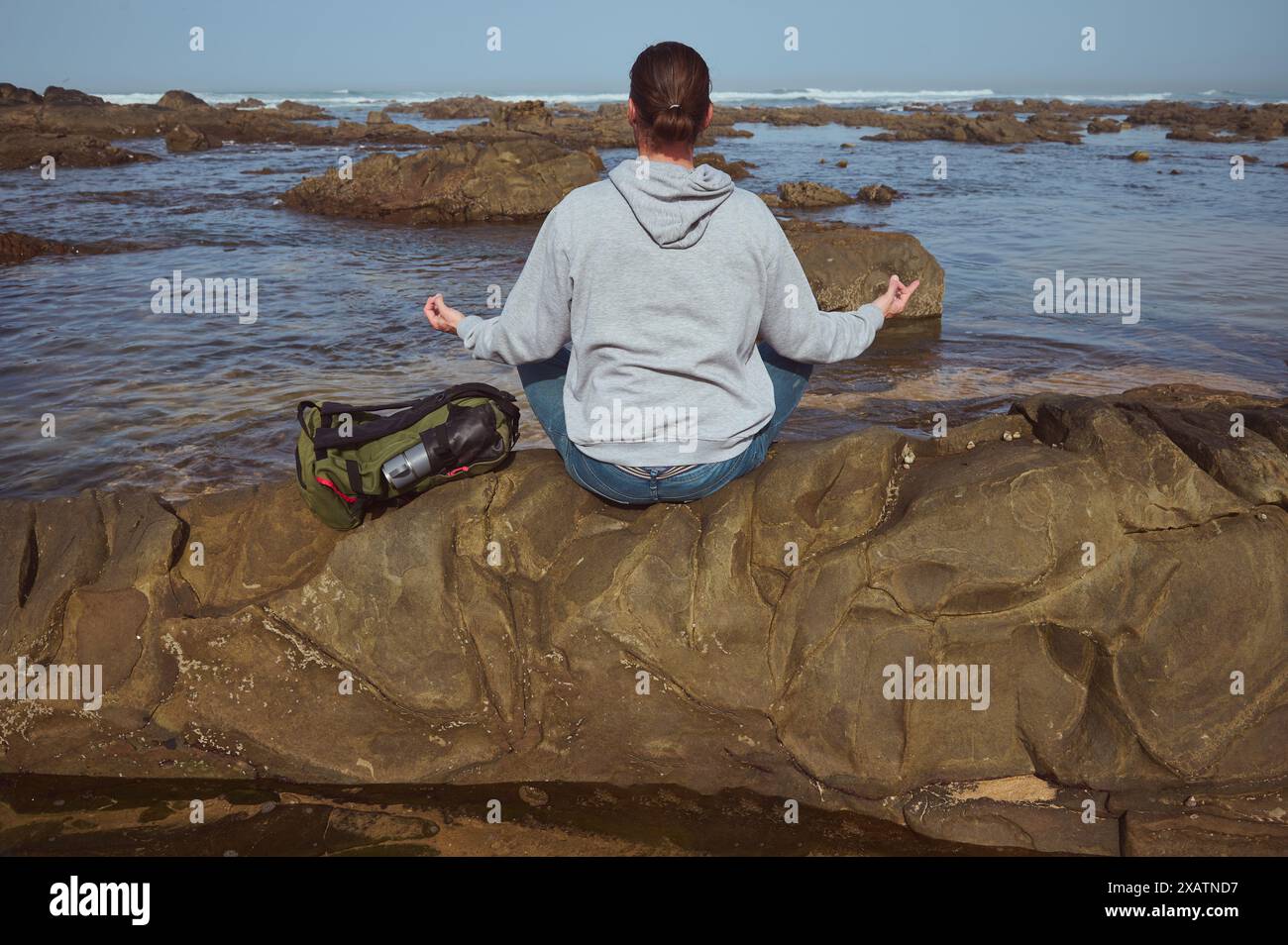 Una persona siede in posa meditativa sulla costa rocciosa, di fronte all'oceano. L'ambiente tranquillo mette in risalto la tranquillità della natura, con le onde che si avvicinano dolcemente Foto Stock