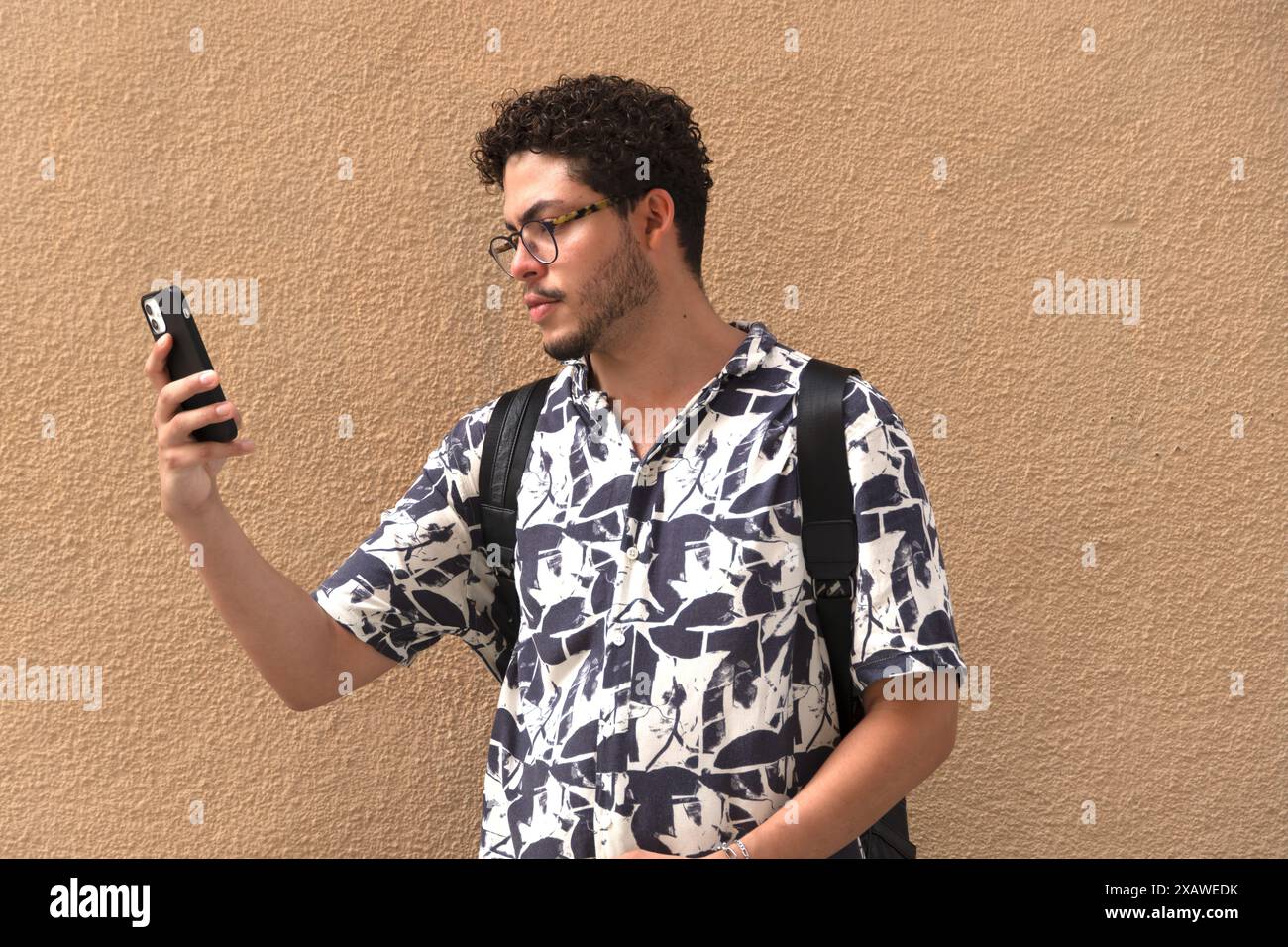 Il giovane moderno fa selfie in città. Capelli ricci, occhiali, camicia stampata, moda urbana Foto Stock