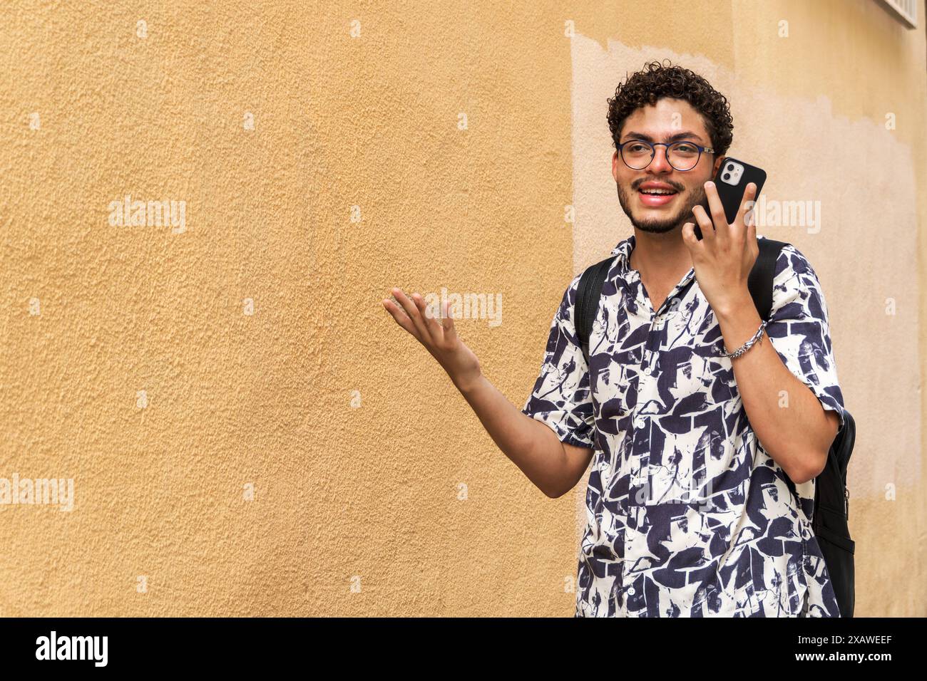 Giovane uomo moderno con occhiali e zaino, che utilizza smartphone, sorridente e gestuale. Comunicazione, tecnologia, moda urbana, stile di vita Foto Stock