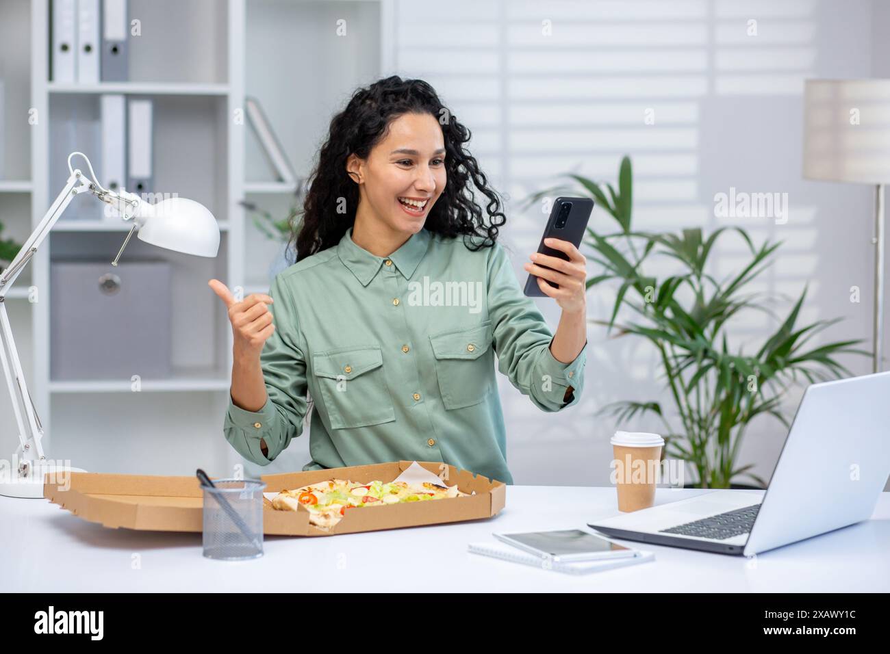 Una donna allegra che pranza alla sua scrivania mentre videochiama con lo smartphone, mostrando i pollici in un moderno ufficio con laptop e caffè. Foto Stock