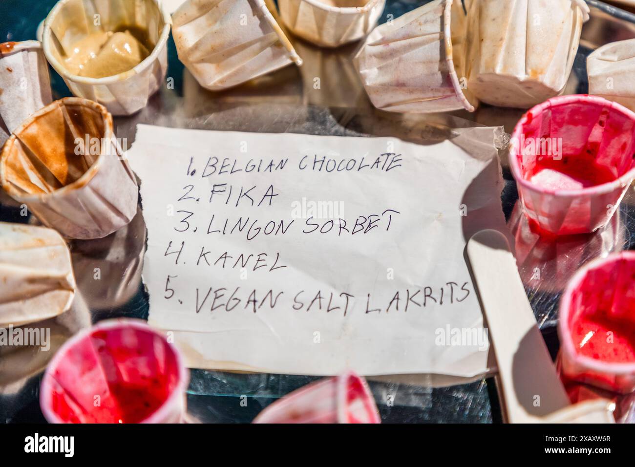 Una nota scritta a mano elenca cinque gusti di gelato Stikki Nikki: cioccolato belga, fika, sorbetto ai mirtilli rossi, kanel (cannella) e liquirizia salata vegana. Ciotole di gelato vuote e parzialmente piene con il sorbetto ai mirtilli rossi sono sparse intorno alla nota. Skanstull, Södermalm, Stoccolma, Contea di Stoccolma, Svezia. Foto Stock