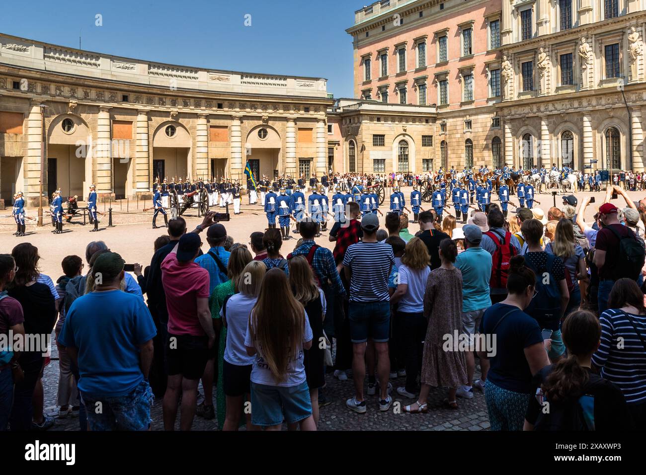 Gli spettatori alzano i loro smartphone per fotografare la guardia schierata davanti alla colonnata curva del Kungliga Slottet a Stoccolma, mentre la banda di cavalleria a cavallo è già visibile in avvicinamento sullo sfondo a destra. Yttre borggården, Stoccolma, Contea di Stoccolma, Svezia. Foto Stock
