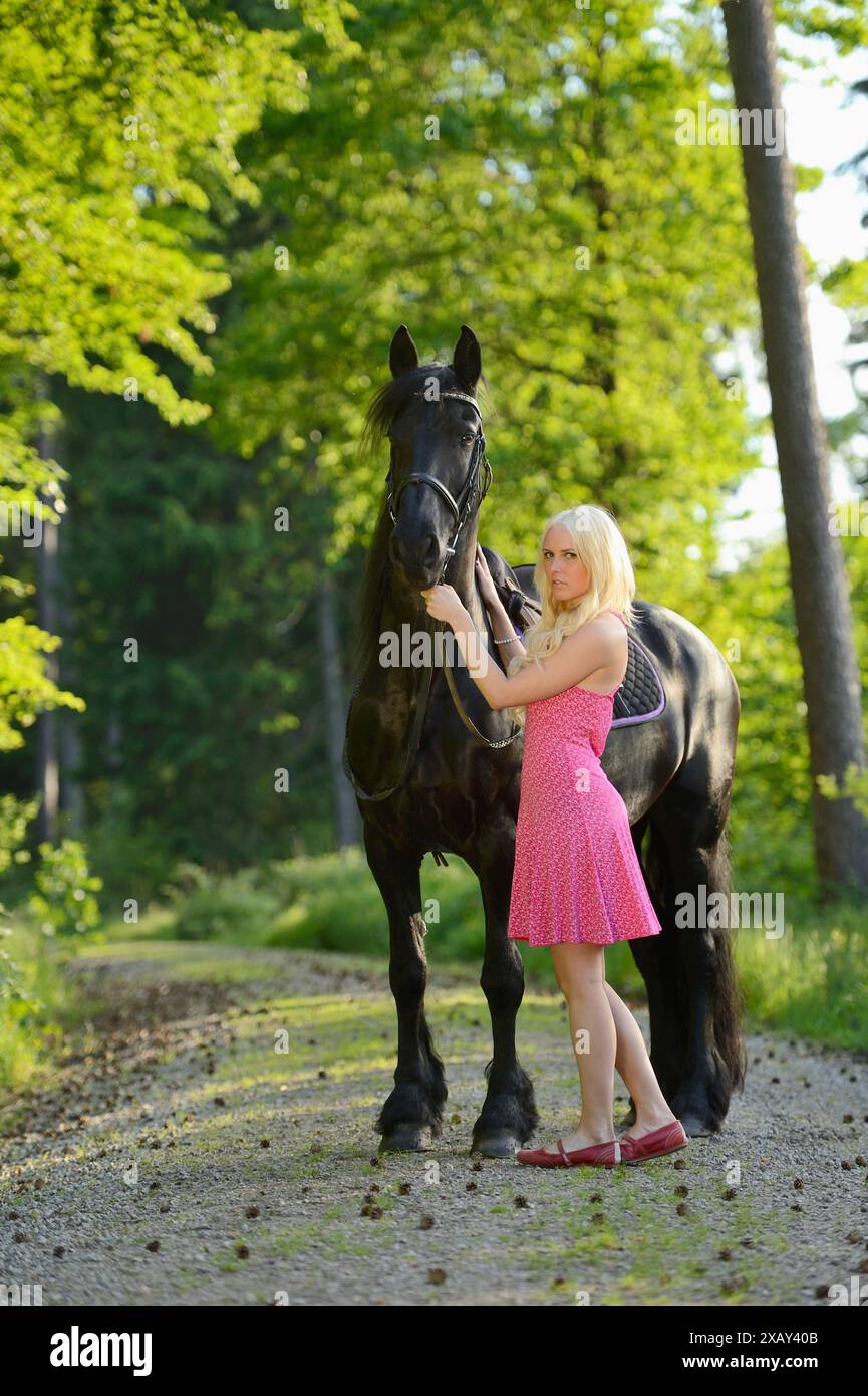 Donna bionda con un abito rosa che tiene in braccio un cavallo frisone nero in un ambiente forestale, la Baviera Foto Stock