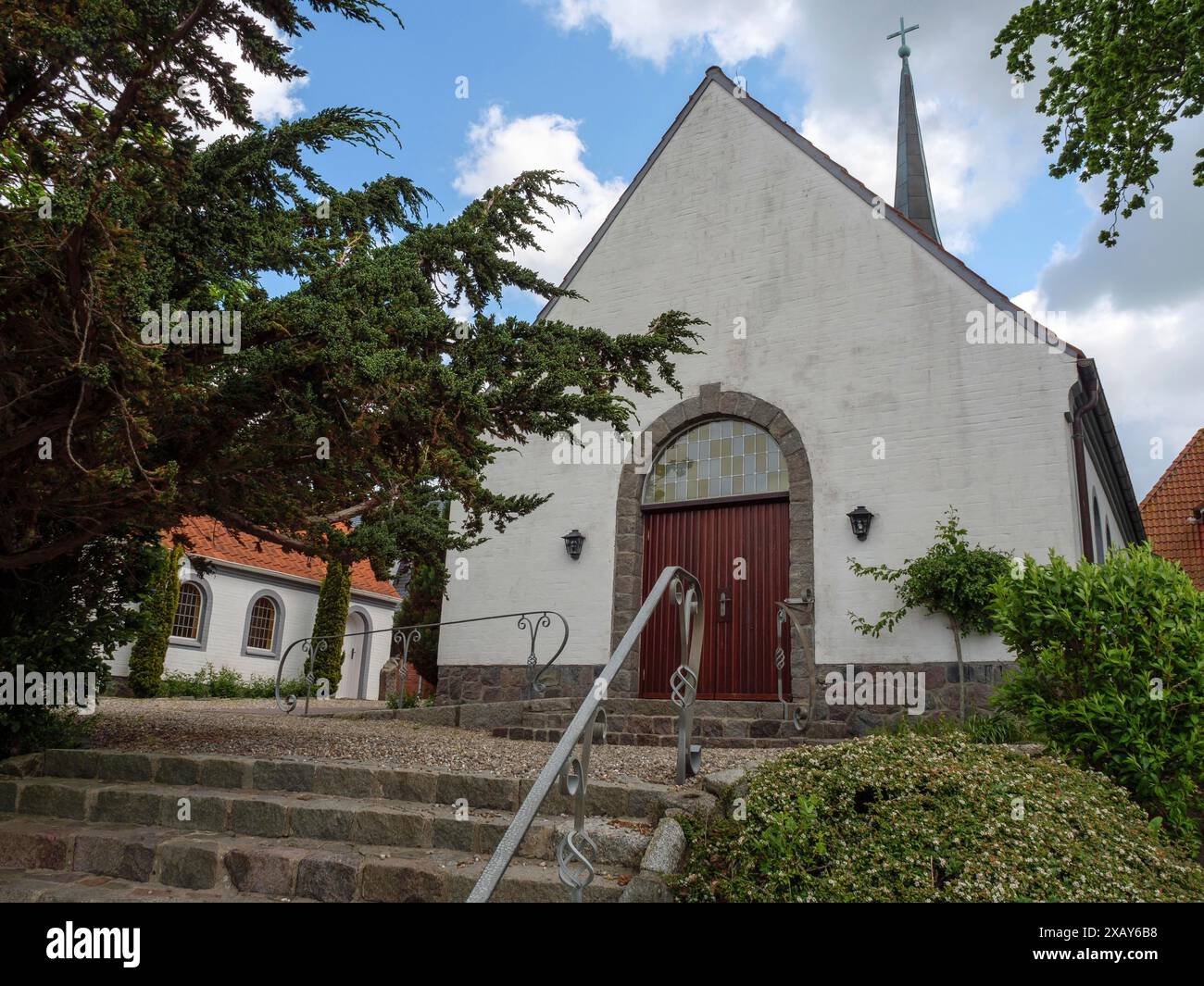 Chiesa bianca con porte rosse, circondata da alberi e una scalinata in pietra sotto un cielo blu, Maasholm, Schleswig-Holstein, Germania Foto Stock