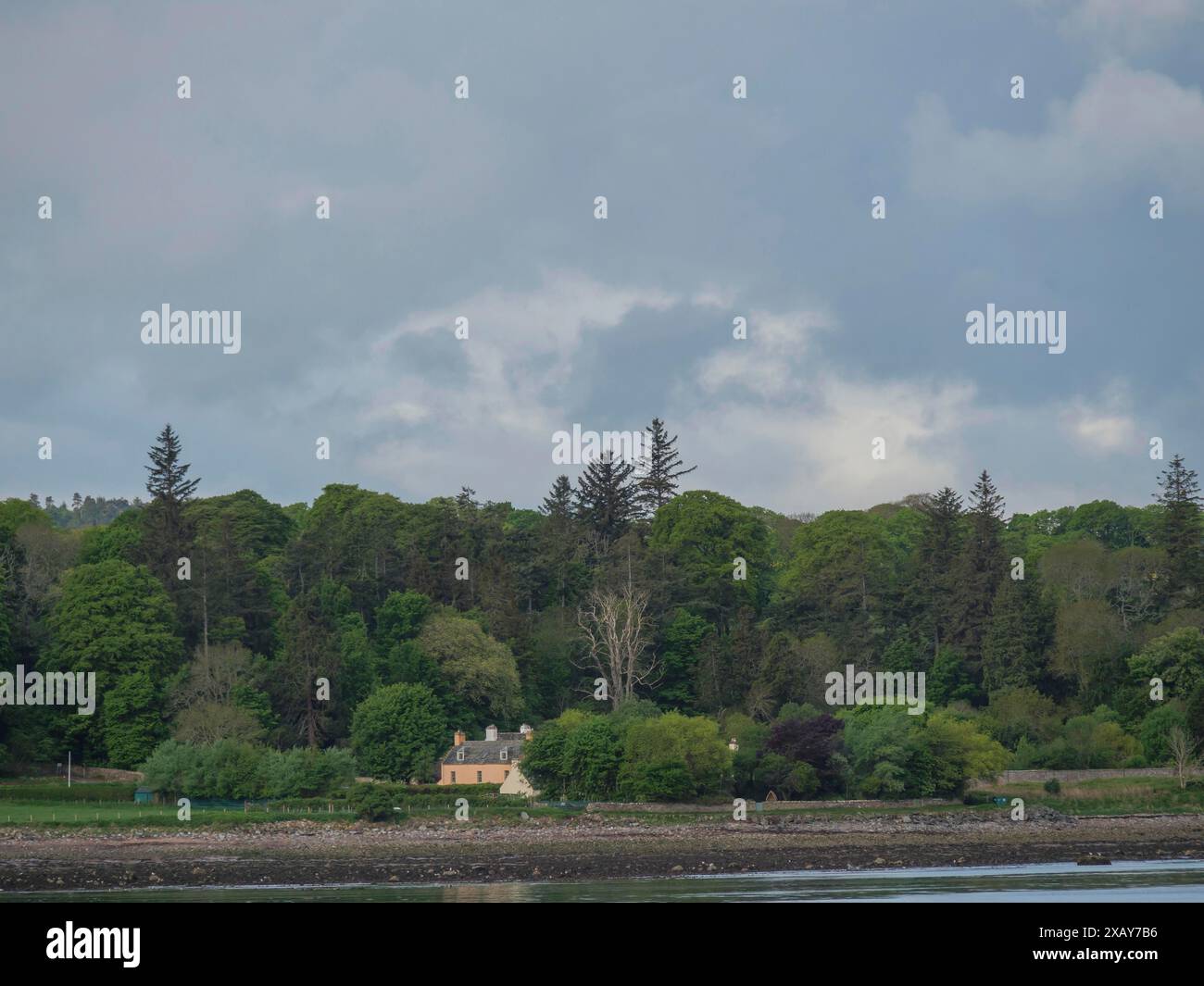 Una piccola casa ai margini di una foresta, circondata da alti alberi e affacciata su una sponda del fiume sotto un cielo nuvoloso, inverness, Scozia, Regno Unito Foto Stock