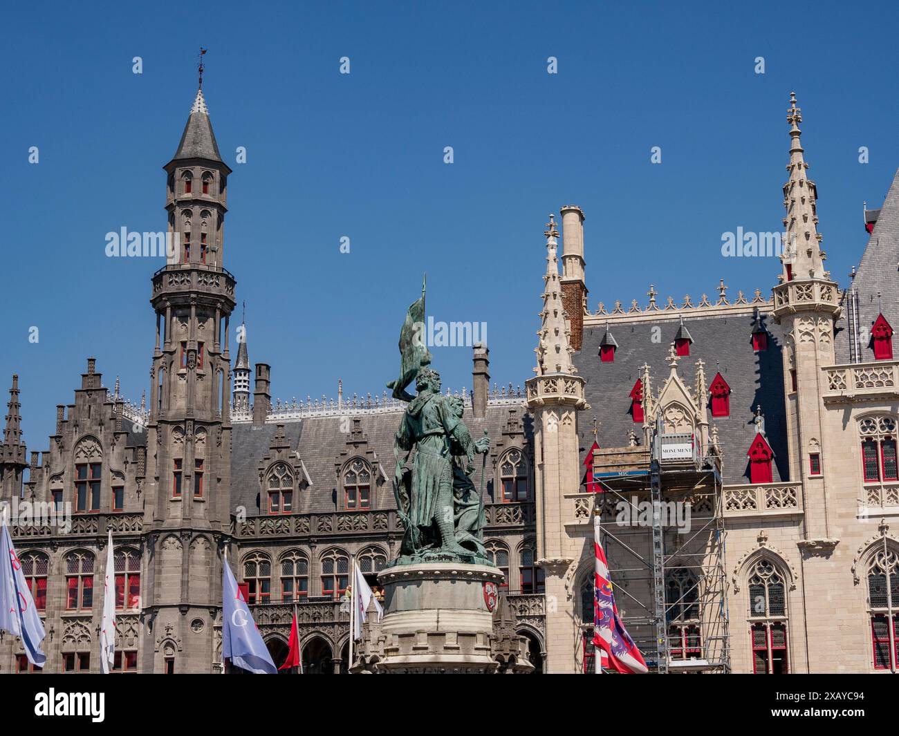 Una statua di fronte a magnifici edifici gotici con bandiere ondulate sotto un cielo azzurro, Bruges, Belgio Foto Stock