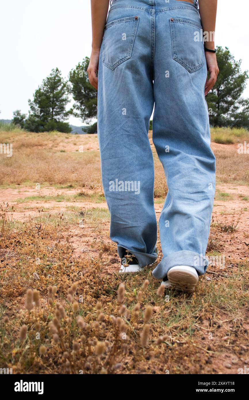 Donna in natura in una passeggiata in estate durante il giorno indossando jeans blu, vivendo uno stile di vita sano, erba verde, alberi e fiori Foto Stock
