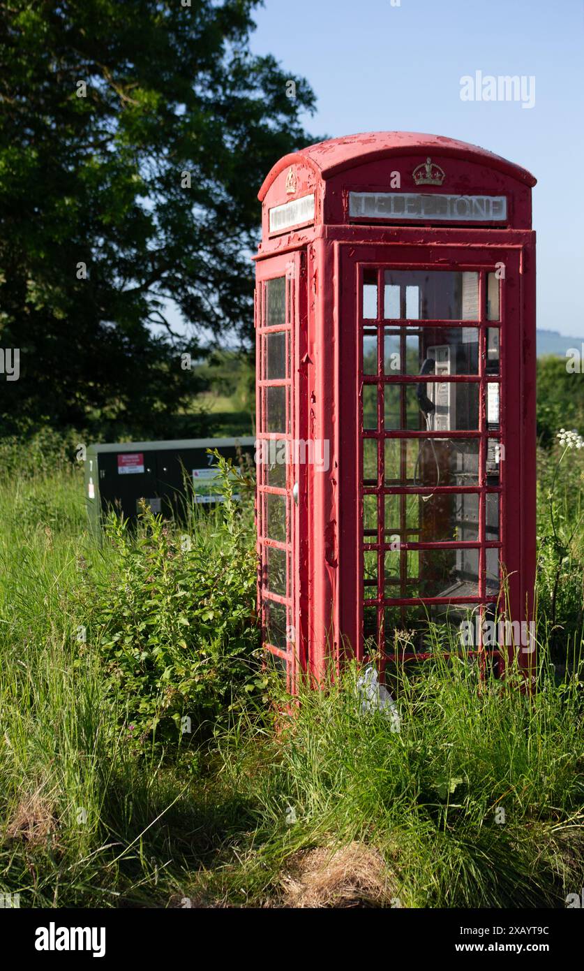 Tradizionale cabina telefonica rossa britannica inutilizzata in un ambiente rurale sovrastato da vegetazione verde e cielo azzurro Foto Stock