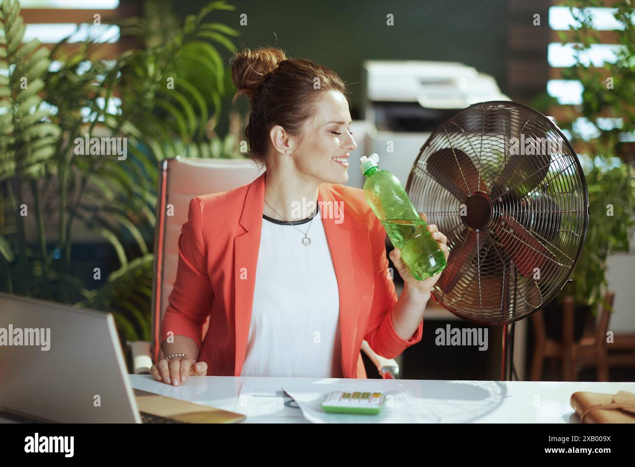 Ambiente di lavoro sostenibile. lavoratrice moderna e felice in un moderno ufficio verde con giacca rossa con bottiglia d'acqua, ventilatore elettrico e laptop. Foto Stock