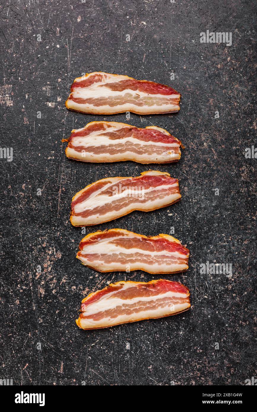 Immagine ravvicinata di cinque strisce di bacon crude disposte su una superficie nera e ruvida. Vista dall'alto. Foto Stock