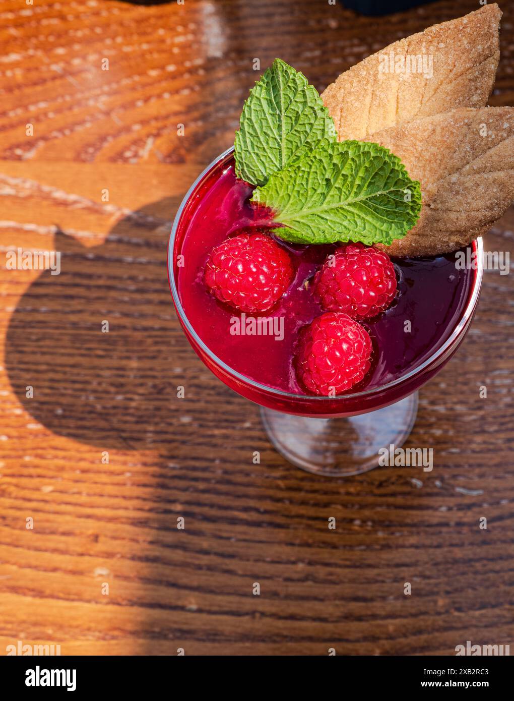 Un bicchiere di mousse di lamponi condita con frutti di bosco freschi, menta e biscotti, una presentazione illuminata dal sole su una superficie in legno. Foto Stock
