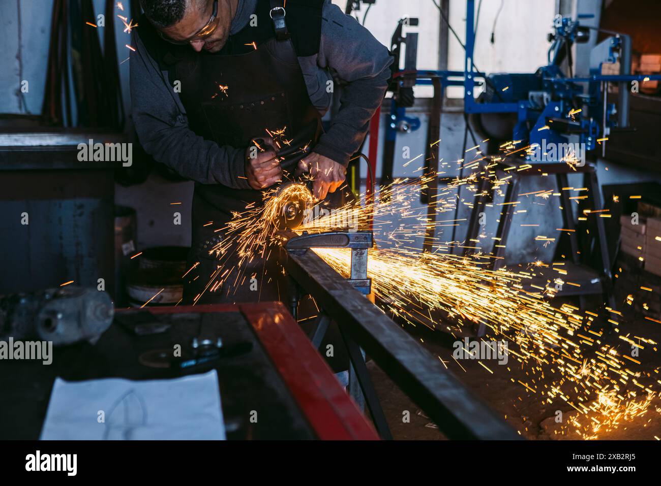 Un fabbro corto e irriconoscibile macina il metallo su un'incudine, con scintille luminose che volano intorno a un laboratorio poco illuminato. Foto Stock