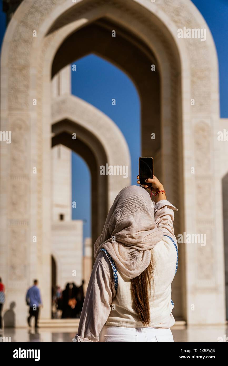 Una donna anonima con un abbigliamento modesto scatta una foto con il suo smartphone, incorniciata da eleganti arcate dell'Oman, catturando l'essenza del suo viaggio culturale Foto Stock