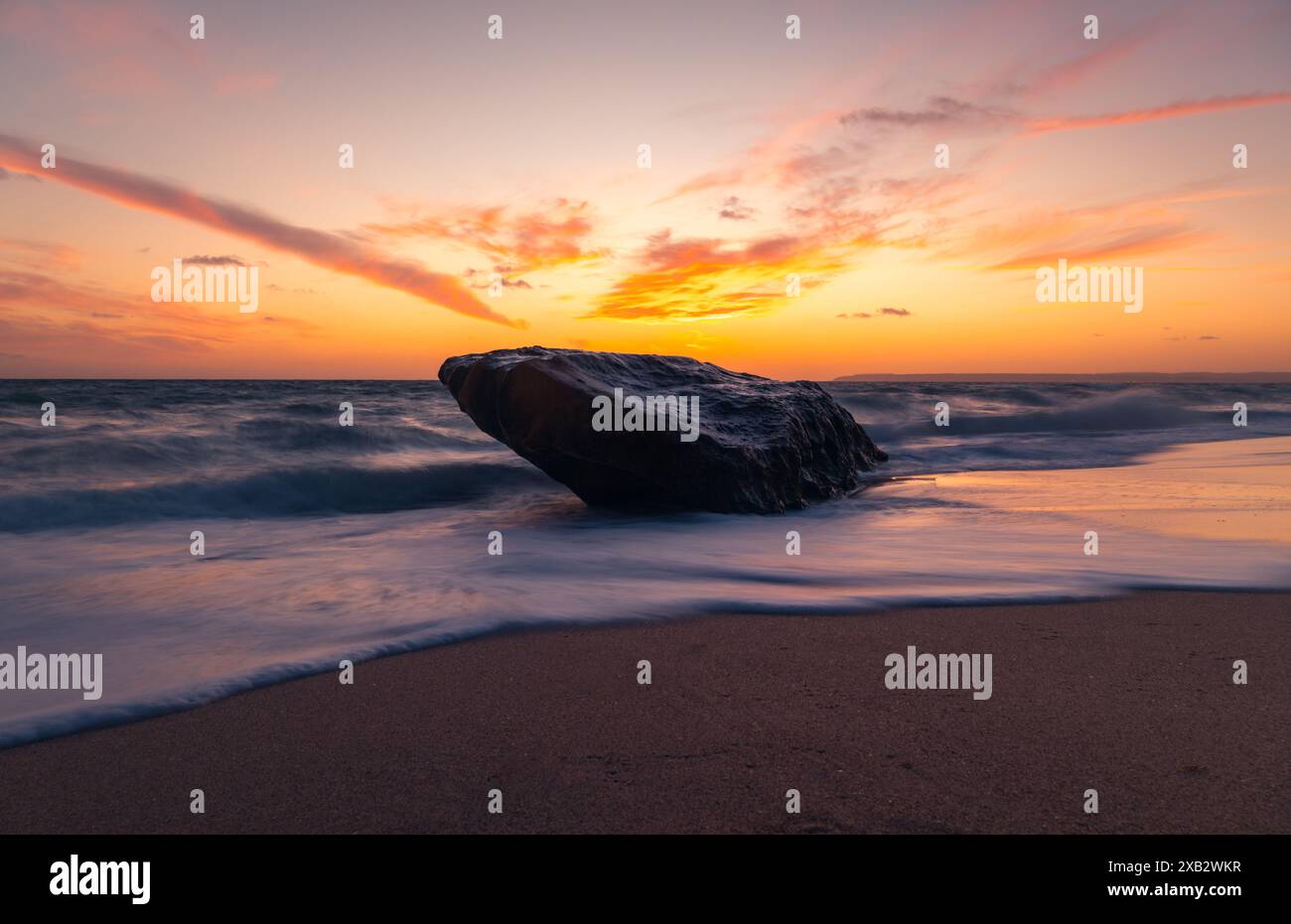 Le vivaci tonalità del tramonto dipingono il cielo sopra Atlanterra Beach mentre le onde accarezzano un masso solitario sulla spiaggia sabbiosa. Foto Stock