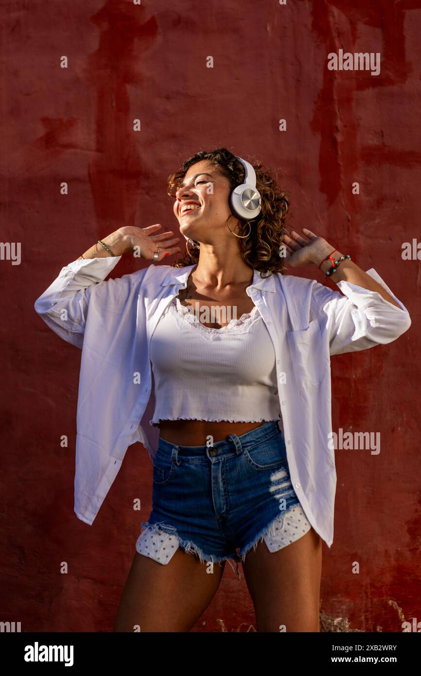 Una giovane donna felice che indossa una camicia bianca e un paio di pantaloncini in denim ama ascoltare la musica con le cuffie su un vivace sfondo rosso Foto Stock