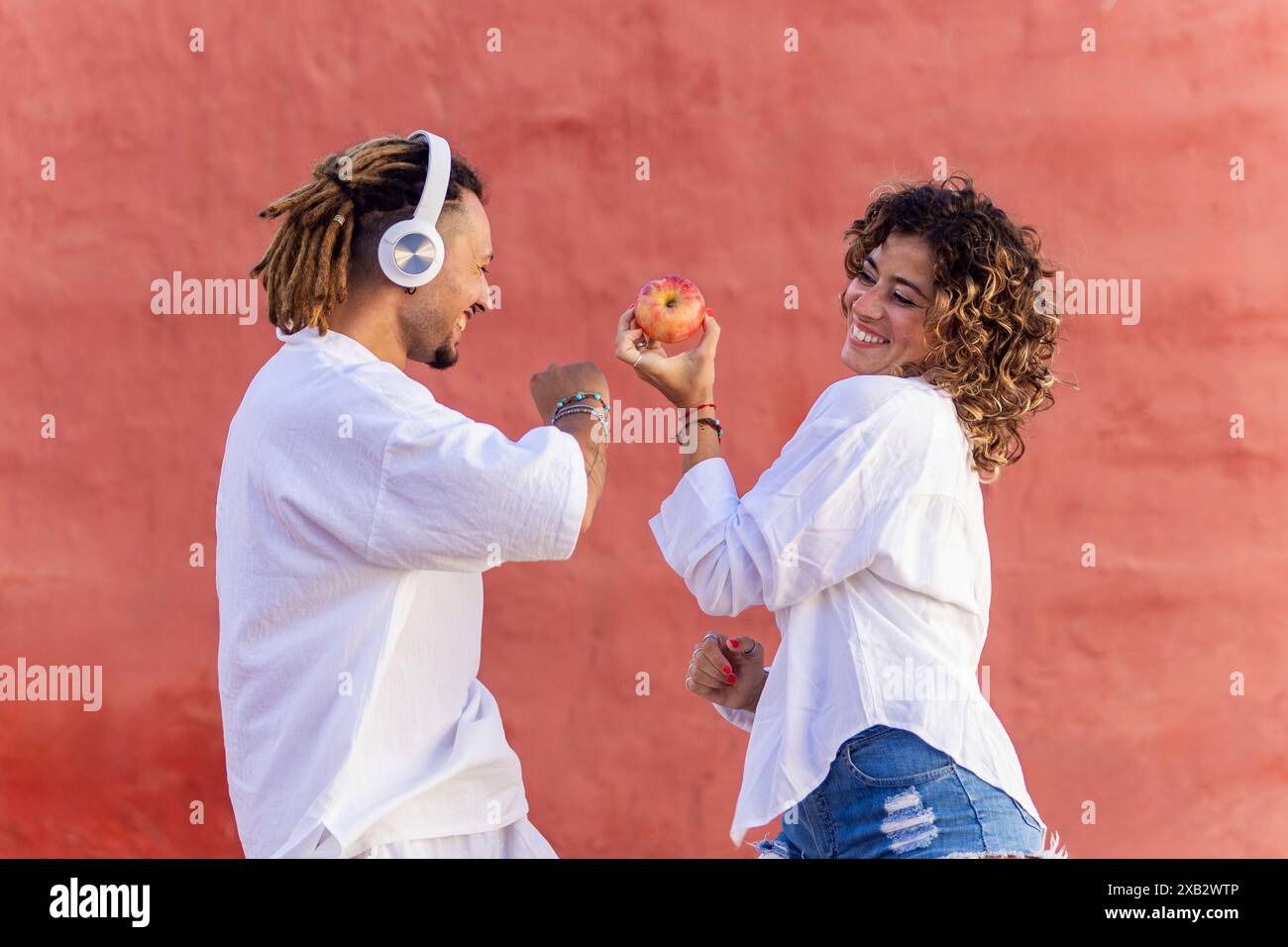 Un uomo e una donna gioiosi condividono un momento di gioco all'aperto, tenendo una mela con sorrisi, vestita in casual top bianchi, contro un muro rosso Foto Stock