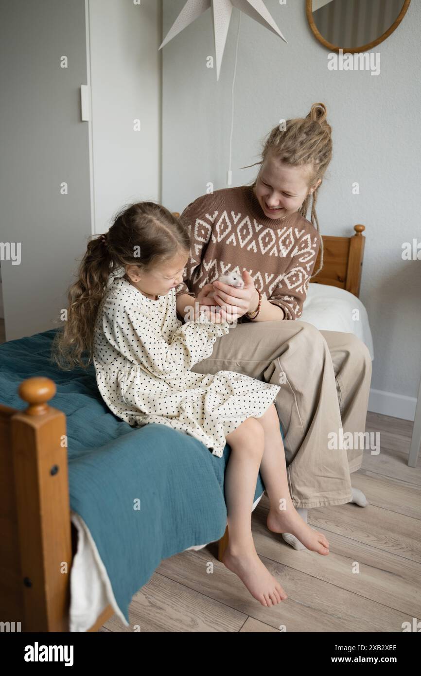 Una madre e una figlia sorridenti, comodamente sedute su un letto, condividono un momento di tenerezza tenendo delicatamente in mano un criceto. Entrambi guardano in basso la loro F. Foto Stock