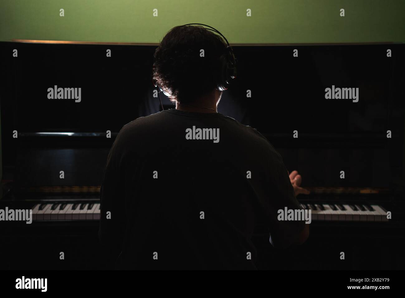 Un uomo in una stanza buia con un pianoforte a coda, visto da dietro, indossa le cuffie mentre coinvolge intensamente la musica. L'immagine cattura un momento di artisti Foto Stock