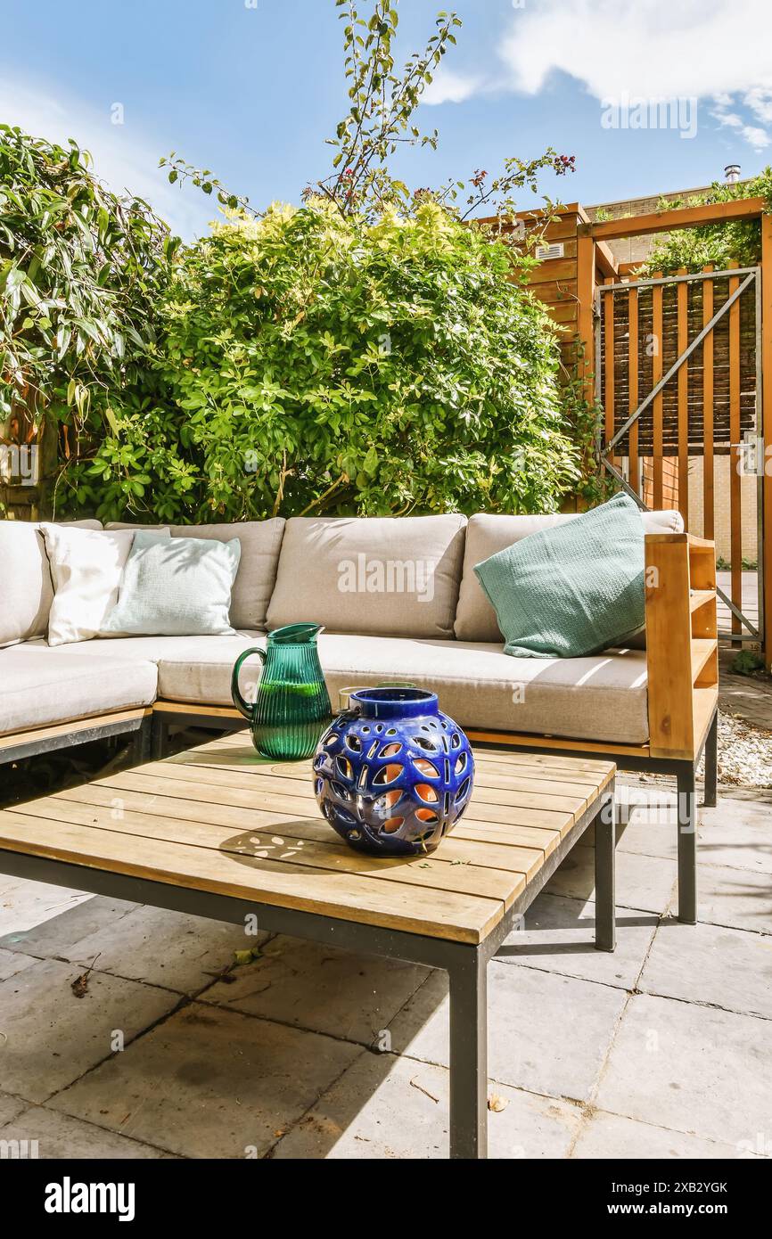 Un'invitante area patio esterna con un moderno divano angolare, tavolino da caffè in legno e vasi decorativi in vetro blu, circondati da una vegetazione lussureggiante. Foto Stock