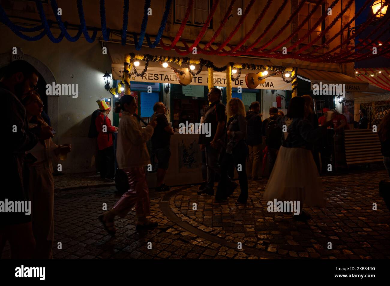 Le persone si riuniscono intorno a una bancarella di cibo festivo ad Alfama durante le popolari festività dei Santi, con decorazioni scintillanti e luci che si aggiungono alla scena Foto Stock