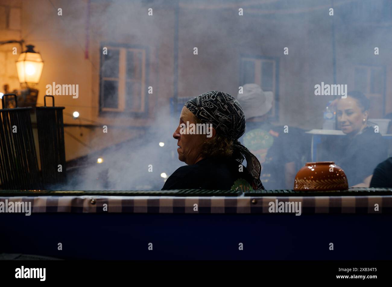 Donna con un velo a motivi geometrici che lavora all'interno di una bancarella di cibo durante le festività di alfama, circondata da fumo, colleghi sullo sfondo Foto Stock