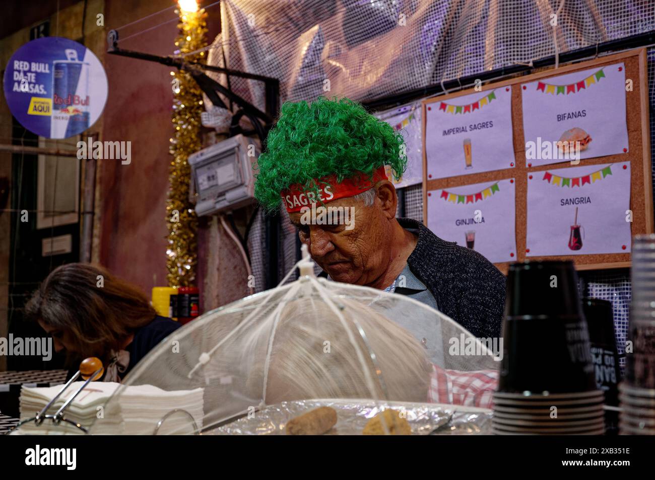 Un uomo maturo con parrucca verde intensamente concentrato all'interno di un chiosco di cibo durante le celebrazioni dei santi di alfama, un colorato menu illustrato dietro di lui Foto Stock